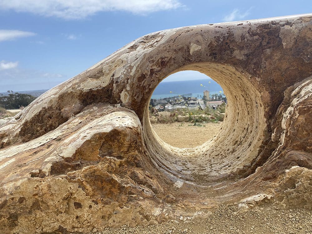A natural rock formation with a circular hole frames a view of the coastal cityscape and the ocean under a blue sky, capturing the serene beauty as if nature itself were entering an In Bloom Contest.