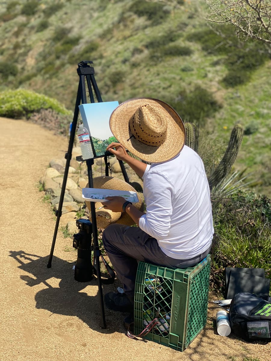 A person wearing a straw hat sits on a plastic crate and paints a landscape for the In Bloom Contest, working at an easel along a dirt path in a hilly, green outdoor setting.