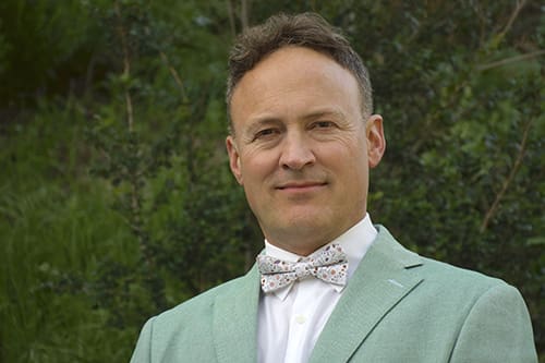 A man wearing a light green suit jacket, white shirt, and patterned bow tie stands outdoors in front of greenery, looking at the camera.
