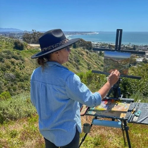 A vibrant woman painting outdoors on a hillside overlooking the ocean at Ventura Botanical Gardens.