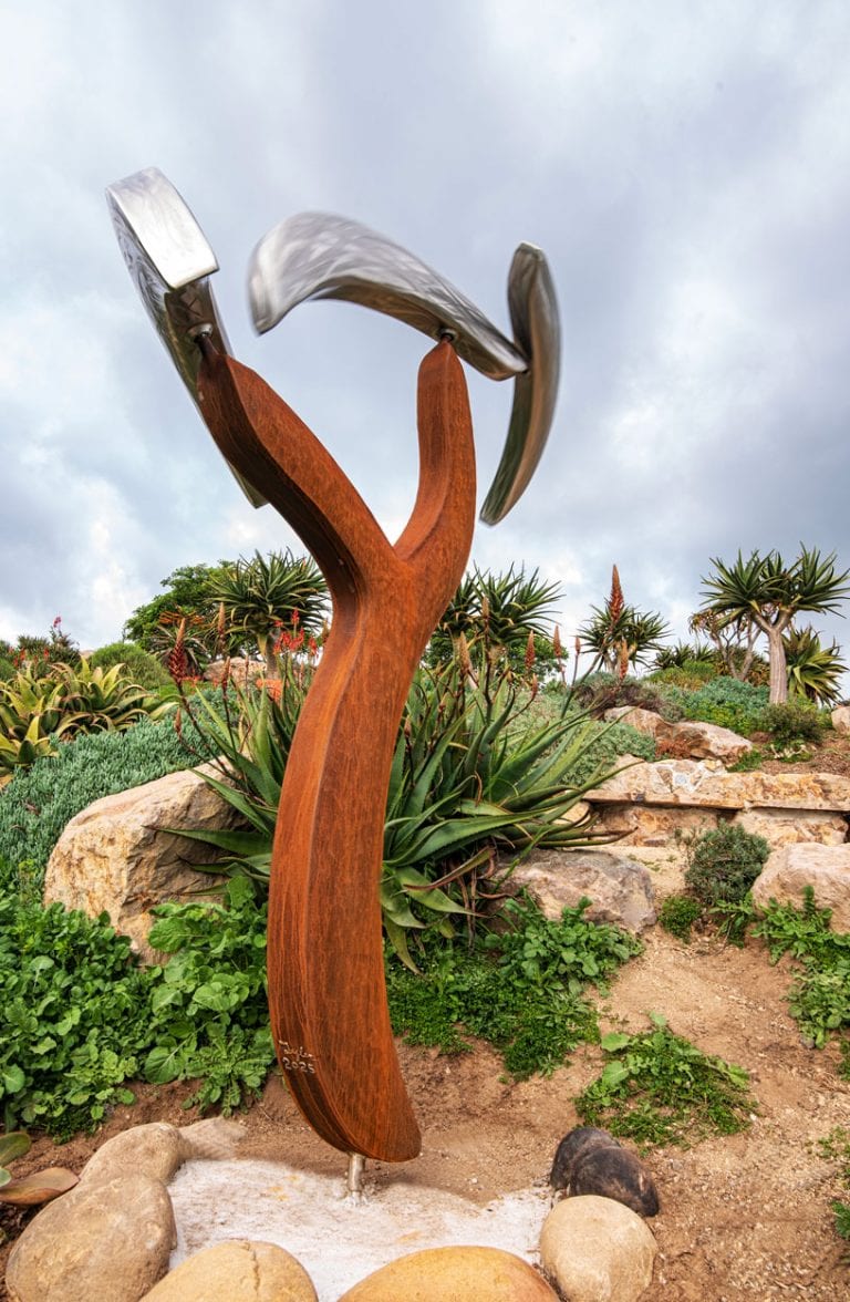 Ventura Botanical Gardens sculpture with metal and wood elements, lush plants, and cloudy sky.